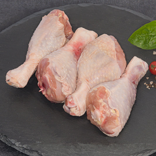 Four raw chicken drumsticks arranged on a dark slate platter with a small green leaf and cherry tomato garnish.