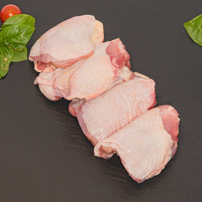 Four raw chicken thighs arranged diagonally on a dark slate plate, garnished with fresh green basil leaves and a small cherry tomato in the corners.