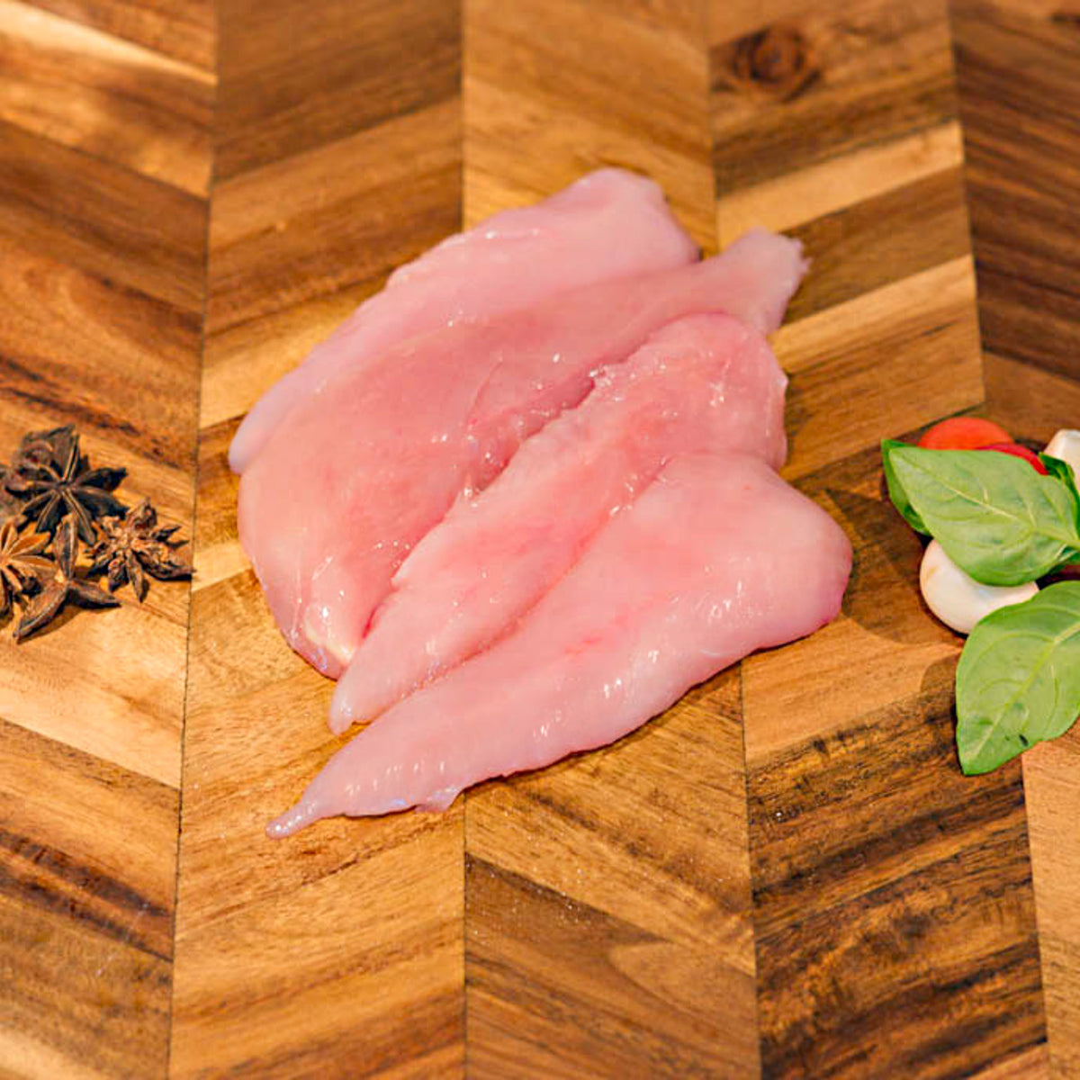 Four raw chicken tenderloins arranged on a wooden cutting board with star anise and basil leaves as props.